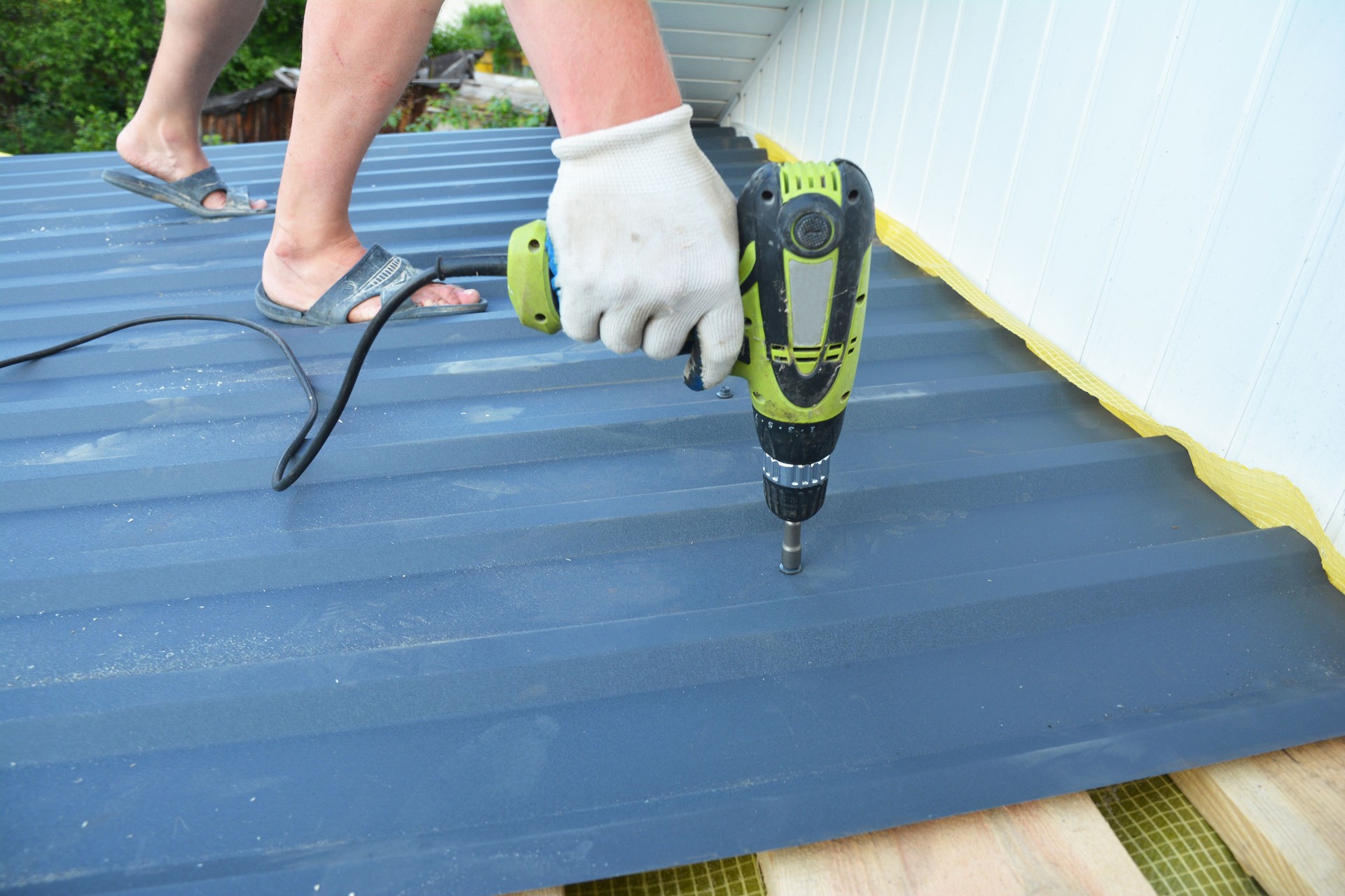 Roofing contractor laying metal roof sheet on the flat house rooftop with power drill during house roofing construction repair.